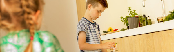 op voorgrond een meisje met vlechten en een groene jurk, gezien vanaf de achterkant. Rechts van haar een kleine jongen met zwarte broek en gestreepts short. In zjn linker hand heeft hij een blikje en in zijn rechter hand een verpakking die hij in een open vuilnisbak gooit