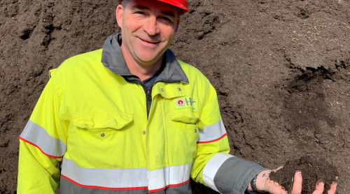Bedrijfsleider staat met veiligheidskleding aan voor een compostberg met compost in zijn hand