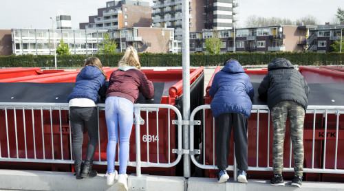 4 kinderen kijken vanaf een balustrade op het afvalbrengstation naar containers