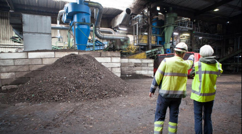 Twee mensen staan in de composteringshal in Middenmeer een van hen wijst naar een installatie en op de voorgrond ligt een hoop compost