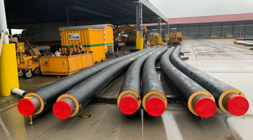 zes warmtebuizen met een rode kop liggen op een bouwterrein. Deze buizen zijn gebogen. De buizen worden gebruikt om het warmtenet aan te leggen. Hierdoor stroomt na de aanleg warm water.