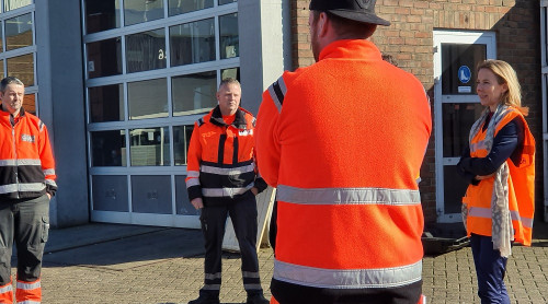 minister Van Veldhoven staat rechts op de foto. OP de rug links van haar staat een chauffeur. Tegenover hem staan nog twee chauffeurs. Op de achtergrond zie je een gebouw