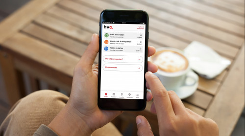 Man houdt mobiel vast en daarop zie je de HVC afval app. Op de achtergrond staat een tafel met daarop een kop cappucino