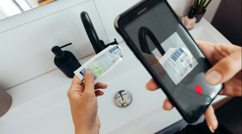 Vrouw staat in de badkamer en houdt in linkerhand een lege tandpastatube vast en in rechterhand haar telefoon. Op scherm van telefoon zie je de barcode scannen-functie geopend van de afvalwijzer uit de HVC afval-app.
