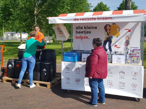 Een inwoner uit Emmeloord levert iets in bij de recyclekraam een afvalcoach van HVC staat er bij en een vrouw staat bij de recyclekraam te kijken