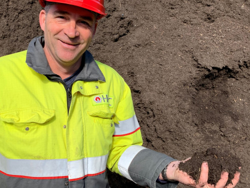 Bedrijfsleider staat met veiligheidskleding aan voor een compostberg met compost in zijn hand