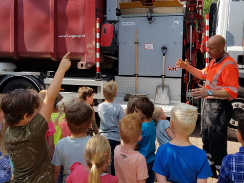 Groep kinderen bij vrachtwagen waar chauffeur uitleg geeft terwijl een van de kinderen een vinger opsteekt