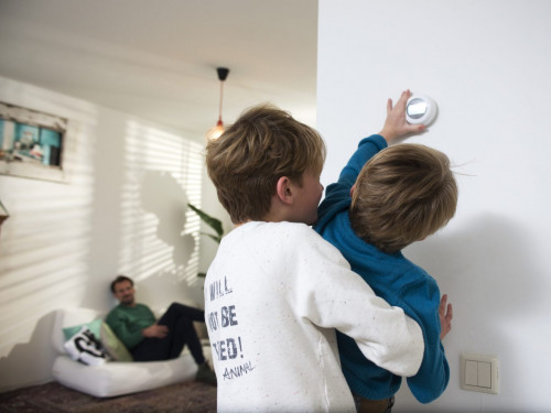 Spelende kinderen in huis draaien aan de muur thermostaat. Vader kijkt vanuit de achtergrond toe. 