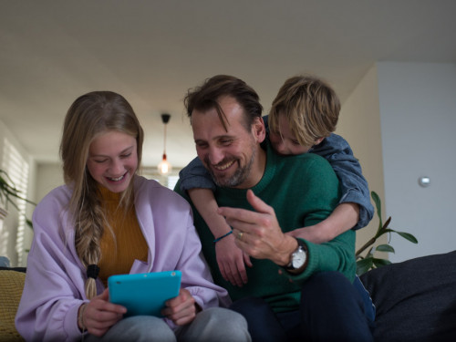 vader en 2 kinderen zitten op de bank en kijken lachend naar een tablet. 