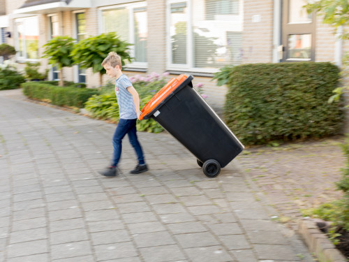 jongen heeft achter zich een bak vast met een oranje deksel. Hij loopt op de stoep voor een huis.