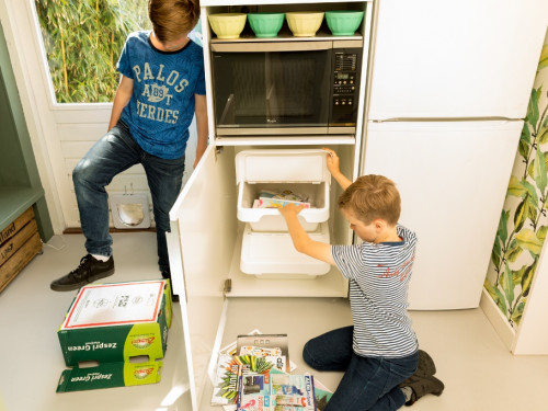 Twee jongens staan in de keuken het oud papier op te ruimen. Een van de jongens stampt een doos plat. De andere jongen zit op de grond en stopt oud papier in een papierkrat die in een keukenkast staat.