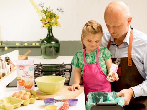 Man rechts en kleine meisje links. Man houdt bakje vast. En meisje doet een drinkpakje erin. Links een tafel met daarop een open doos eieren en een open pak mix voor cupcakes. Ook op de tagel een lege schaal en vier vormen voor cupcakes.