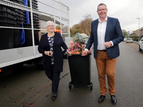 Rechts staat de wethouder, in het midden een plasticbak met daarop een bos bloemen. Links van de bak staat een inwoner. Daarnaast een auto met daarin gestapelde lege bakken.