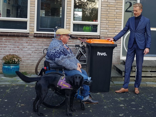 Wethouder staat rechts van bak met oranje deksel. Links zit een man in een rolstoel. Naast hem staat een aangelijnde hond. Ze staan aan de voorkant van een woning.