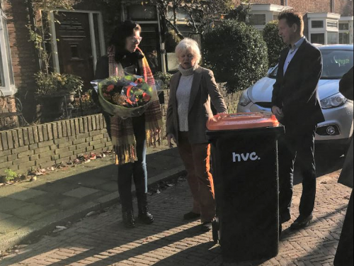 Rechts staat de directeur HVC, links van hem een bak met oranje deksel. Links van de bak staat een mevrouw. Daarnaast staat een mevrouw met een bos bloemen. Ze staan allen op de weg. Op de achtergrond rechts zie je een geparkeerde auto. Op de achtergrond woonhuizen met een tuin
