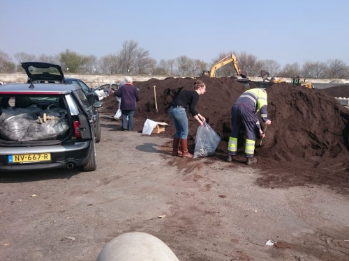 Drie personen staan tijdens compostdag compost te scheppen van een grote berg ine en zak te scheppen naast een auto met in de achterbak al een aantal volle plastic zakken compost