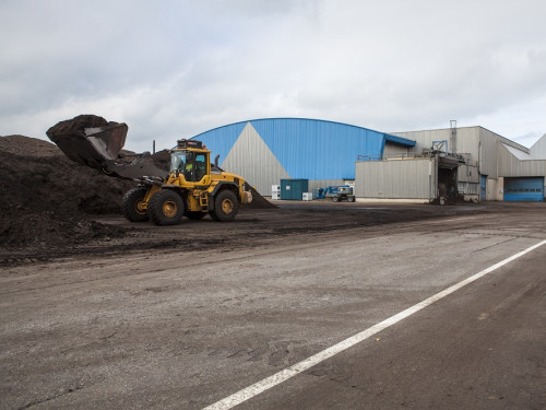 Weg met asfalt. links in de hoek een heftruck bij een hoop compost. Op de achtergrond de blauwe composteerhallen van de composteringsinstallatie in Purmerend
