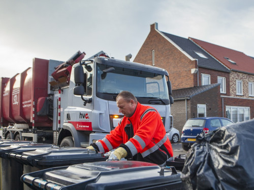 Op de voorgrond staan drie grijze bakken. Een chauffeur van HVC pakt met twee handen de tweede bak vast. Op de achtergrond links zie je een zijlader van HVC. Rechts op de achtergrond zie je een blauwe auto en huizen.