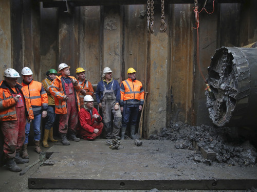 De foto is gemaakt in een bouwput. Op de foto zie je links werkmannen staan, rechts zie je de boor die zich een weg onder spoor door heeft geboord. De boor heeft een diameter die groter is dan de werkmannen die er naar kijken.