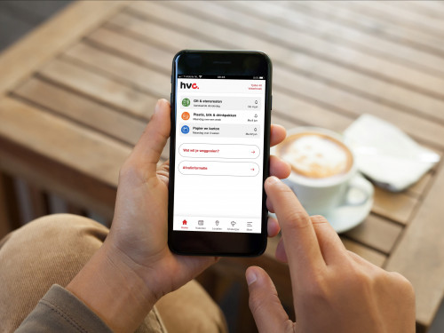 Man houdt mobiel vast en daarop zie je de HVC afval app. Op de achtergrond staat een tafel met daarop een kop cappucino