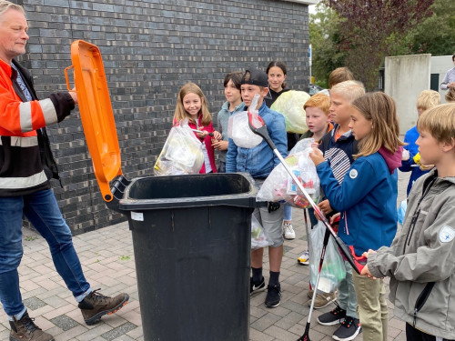 kinderen staan om bak emt oranje deksel. In hun handen hebben ze zakken met afval en afvalprikkers