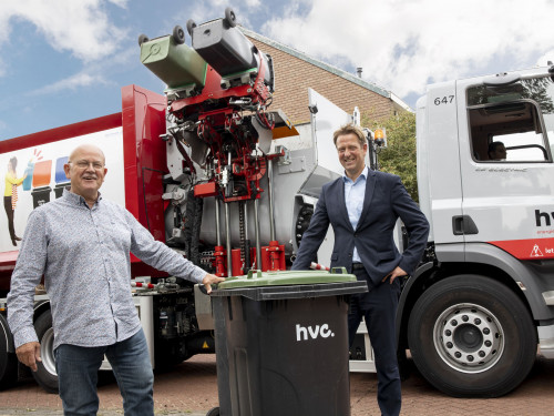 Wethouder Floor Bal en HVC directeur Gertjan de Waard staan voor de elektrische inzamelwagen terwijl ze een groene bak vasthouden.