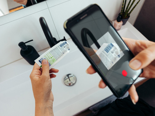 Vrouw staat in de badkamer en houdt in linkerhand een lege tandpastatube vast en in rechterhand haar telefoon. Op scherm van telefoon zie je de barcode scannen-functie geopend van de afvalwijzer uit de HVC afval-app.